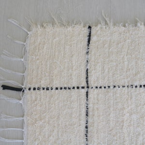 May include: A white rug with black lines forming a grid pattern. The rug has fringe on all sides.