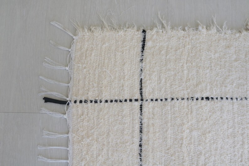 May include: A white rug with black lines forming a grid pattern. The rug has fringe on all sides.