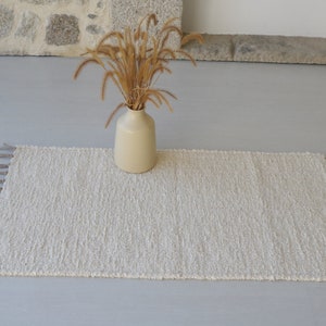 Puede incluir: Una alfombra blanca y esponjosa con flecos en los bordes. La alfombra est&aacute; colocada sobre un suelo gris claro con un jarr&oacute;n de hierba seca en el centro.
