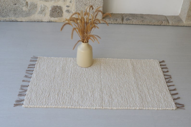 Puede incluir: Una alfombra blanca y esponjosa con flecos en los bordes. La alfombra est&aacute; colocada sobre un suelo gris claro con un jarr&oacute;n de hierba seca en el centro.