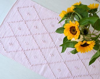 Può includere: Runner da tavolo rosa chiaro con motivo a rombi e piccoli dettagli floreali. Un vaso di girasoli giallo brillante con foglie verdi è posto a destra. Un elemento decorativo per la tavola.