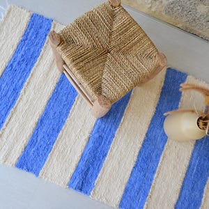 Op de afbeelding: Een rechthoekig geweven vloerkleed met afwisselende verticale strepen in crème en felblauw, afgewerkt met korte franjes aan de uiteinden. Een kleine vierkante houten kruk met een gevlochten zitting en een beige vaas met gedroogde grassen staan erop.