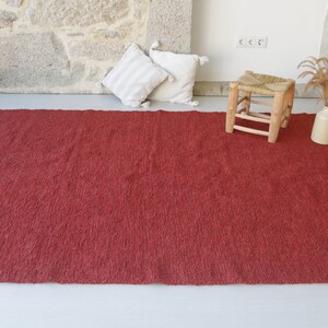 May include: A red, textured rug with a subtle sheen. The rug is laid out on a white floor with a wooden stool and a vase of dried flowers in the background.