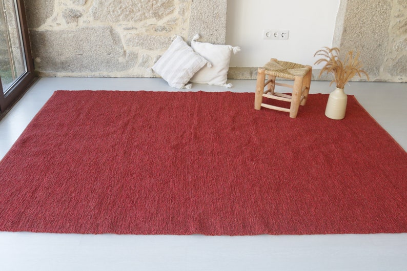 May include: A red, textured rug with a subtle sheen. The rug is laid out on a white floor with a wooden stool and a vase of dried flowers in the background.