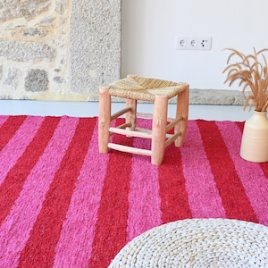 May include: A pink and red striped rug with a woven stool and a vase with dried grass.