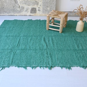 May include: A teal green woven rug with a fringed edge. The rug is laid out on a light grey floor with a wooden stool in the background.