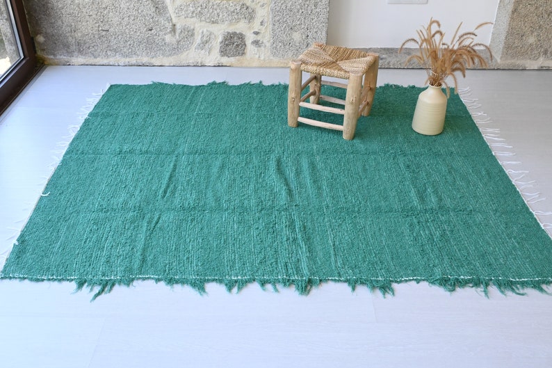 May include: A teal green woven rug with a fringed edge. The rug is laid out on a light grey floor with a wooden stool in the background.