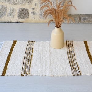 May include: A white and brown striped woven rug with frayed edges. The rug is on a light grey floor with a vase of dried grass in the background.