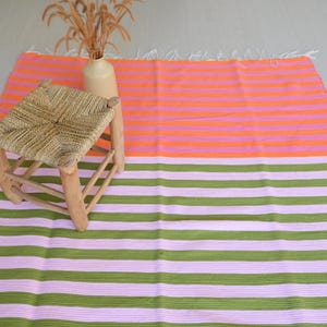 Op de afbeelding: Een rechthoekig vloerkleed met horizontale strepen in twee verschillende kleurblokken. Het bovenste gedeelte heeft afwisselende oranje en roze strepen, terwijl het onderste gedeelte afwisselende olijfgroene en lichtroze strepen heeft.