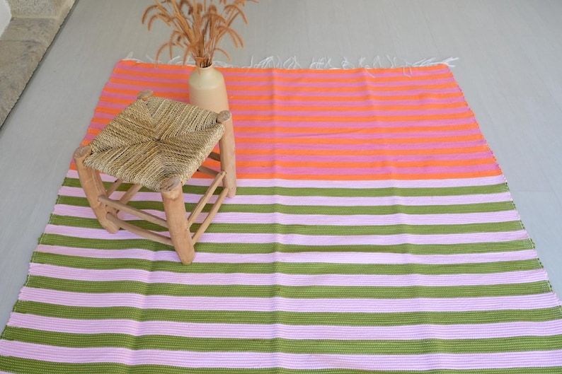 Op de afbeelding: Een rechthoekig vloerkleed met horizontale strepen in twee verschillende kleurblokken. Het bovenste gedeelte heeft afwisselende oranje en roze strepen, terwijl het onderste gedeelte afwisselende olijfgroene en lichtroze strepen heeft.