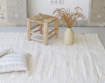 Könnte beinhalten: Ein weißer, flauschiger Teppich mit einem geflochtenen Hocker und einer Vase mit getrocknetem Gras. Zwei weiße Kissen liegen auf dem Teppich.