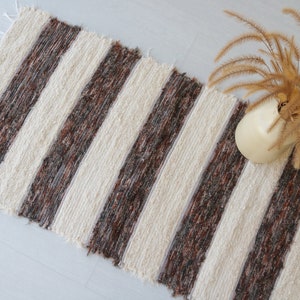 May include: A striped rug featuring alternating bands of brown and cream. The rug has a textured weave and fringed ends. A cream-coloured vase with dried plants is placed on the rug.
