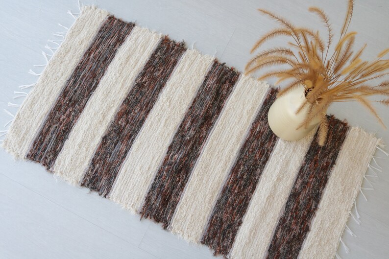 May include: A striped rug featuring alternating bands of brown and cream. The rug has a textured weave and fringed ends. A cream-coloured vase with dried plants is placed on the rug.
