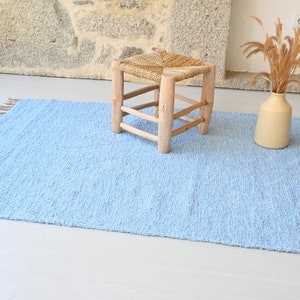 Pu&ograve; includere: Un tappeto blu chiaro con frange sui bordi. Il tappeto &egrave; su un pavimento bianco e uno sgabello di legno con una seduta intrecciata &egrave; sopra il tappeto. Un vaso con erba secca &egrave; sul pavimento accanto al tappeto.