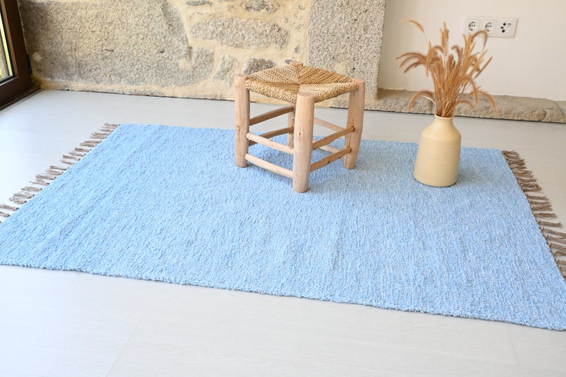 Pu&ograve; includere: Un tappeto blu chiaro con frange sui bordi. Il tappeto &egrave; su un pavimento bianco e uno sgabello di legno con una seduta intrecciata &egrave; sopra il tappeto. Un vaso con erba secca &egrave; sul pavimento accanto al tappeto.