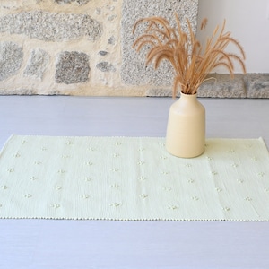 May include: A light green woven rug with a subtle textured pattern. The rug is placed on a light gray floor with a vase of dried grass in the background.