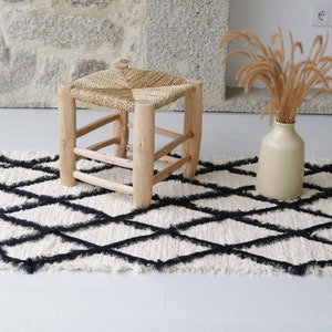 Puede incluir: Una alfombra de patrón de diamantes blancos y negros con un taburete de madera tejido y un jarrón de cerámica beige con hierba seca.