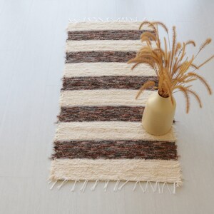 May include: A rectangular, handwoven rug with alternating cream and dark brown stripes. A beige vase holds dried ornamental grass, adding a touch of natural decor. The rug's fringed edges enhance its rustic charm.