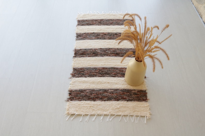May include: A rectangular, handwoven rug with alternating cream and dark brown stripes. A beige vase holds dried ornamental grass, adding a touch of natural decor. The rug's fringed edges enhance its rustic charm.