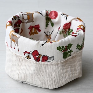 Pode incluir: Uma pequena cesta de tecido com exterior texturizado creme. O interior e a borda superior apresentam uma estampa festiva com Papai Noel, renas e decorações de feriados. A cesta é projetada para armazenamento e decoração.