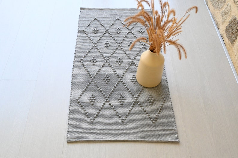 Puede incluir: Una alfombra gris y blanca con un dise&ntilde;o de diamantes. La alfombra est&aacute; hecha de un material texturizado y tiene un brillo sutil. Un jarr&oacute;n con flores secas est&aacute; sentado sobre la alfombra.