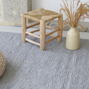 May include: A gray woven rug with fringe on the edges. The rug is on a light wood floor. There is a small wooden stool with a woven seat and a vase with dried grass in the background.