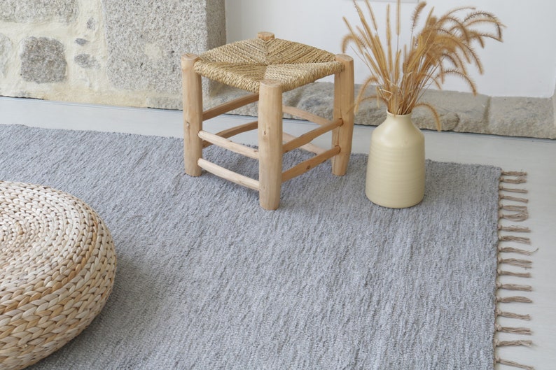May include: A gray woven rug with fringe on the edges. The rug is on a light wood floor. There is a small wooden stool with a woven seat and a vase with dried grass in the background.