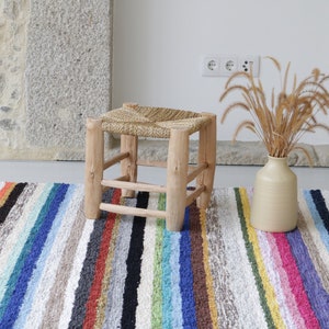 Puede incluir: Un taburete de madera natural con asiento de paja trenzada se encuentra sobre una alfombra de rayas multicolores. Un jarr&oacute;n con hierba seca se encuentra sobre la alfombra.