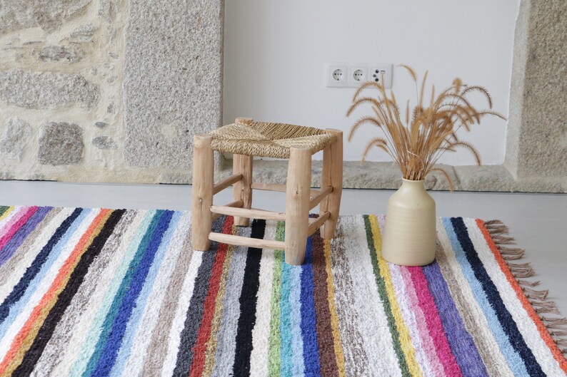 Puede incluir: Un taburete de madera natural con asiento de paja trenzada se encuentra sobre una alfombra de rayas multicolores. Un jarr&oacute;n con hierba seca se encuentra sobre la alfombra.