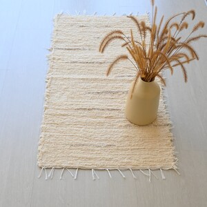 May include: A cream-colored woven rug with frayed edges, featuring a vase with dried grass stems on top. The rug is on a light wood floor.