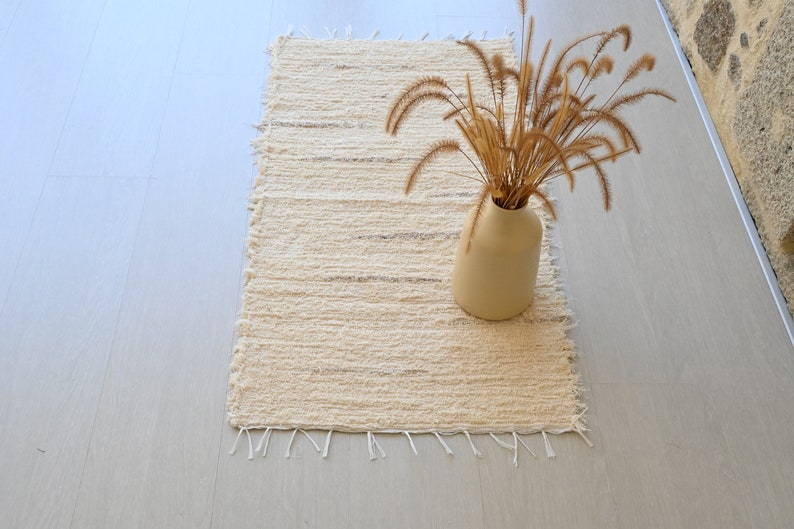 May include: A cream-colored woven rug with frayed edges, featuring a vase with dried grass stems on top. The rug is on a light wood floor.