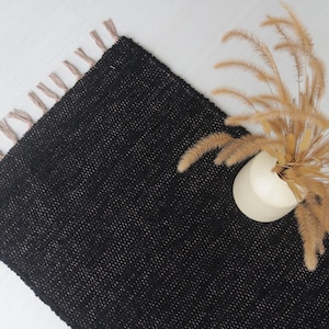 Może przedstawiać: Czarny tkany dywan z frędzlami na krawędziach. Dywan wykonany jest z fakturowanego materiału i ma subtelny połysk. Wazon z suszoną trawą znajduje się na dywanie.