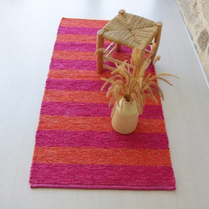 May include: A pink and orange striped rug with a woven stool and a vase of dried flowers on top.