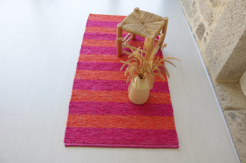 May include: A pink and orange striped rug with a woven stool and a vase of dried flowers on top.