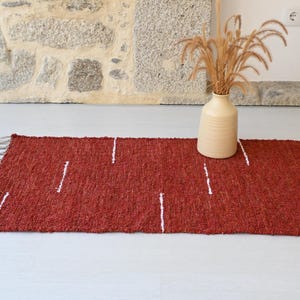 May include: A rectangular, rust-coloured rug with white vertical stripe accents and fringed edges. A beige vase with dried plants sits on the rug. The rug is approximately 91 cm long.