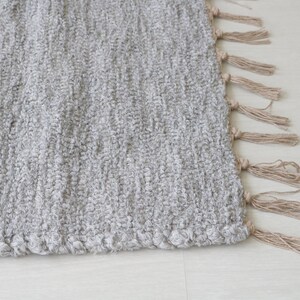 May include: A gray woven rug with a fringe border. The fringe is made of natural fibers and is a light brown color.