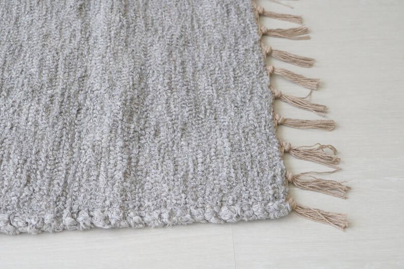 May include: A gray woven rug with a fringe border. The fringe is made of natural fibers and is a light brown color.