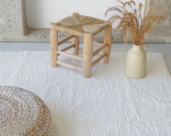 May include: A white woven rug with a natural woven stool and a vase with dried grass.