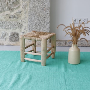 K&ouml;nnte beinhalten: Ein geflochtener Strohhocker mit einem Holzrahmen steht auf einem t&uuml;rkisfarbenen Teppich. Im Hintergrund steht eine Vase mit getrocknetem Gras.