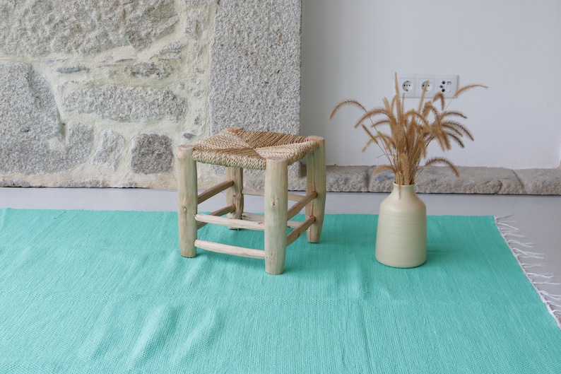 K&ouml;nnte beinhalten: Ein geflochtener Strohhocker mit einem Holzrahmen steht auf einem t&uuml;rkisfarbenen Teppich. Im Hintergrund steht eine Vase mit getrocknetem Gras.