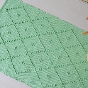 以下が含まれることがあります： 菱形の模様が特徴的な、長方形の明るい緑色のラグ。 各ひし形は、小さな隆起した点で縁取られ、4枚の花びらの花の模様が含まれています。 ラグは織物でできており、明るい色の表面に表示されています。