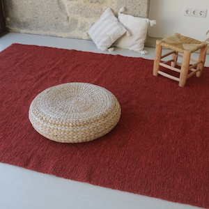 May include: A red woven rug with a round woven ottoman and a wooden stool. The rug is on a white floor.