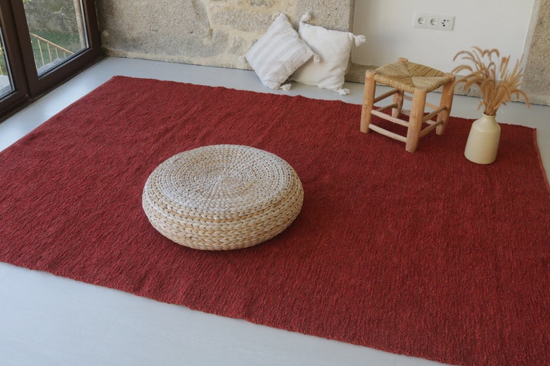 May include: A red woven rug with a round woven ottoman and a wooden stool. The rug is on a white floor.