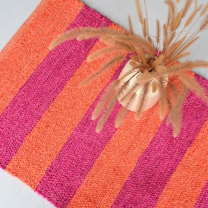 Puede incluir: Una alfombra a rayas verticales en tonos naranja y fucsia. El diseño de la alfombra presenta rayas verticales en negrita. Un pequeño jarrón dorado con hierba ornamental seca se encuentra sobre la alfombra.