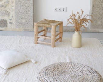 Könnte beinhalten: Ein weißer gewebter Teppich mit einem Holzhocker, einem weißen Kissen und einer runden gewebten Ottomane. Eine Vase mit getrocknetem Gras steht auf dem Teppich.