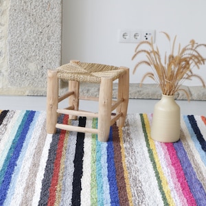 May include: A woven straw stool with a wooden frame sits on a colourful striped rug. The rug has a fringe on the edges and is made of a variety of colours, including red, blue, green, yellow, and white.