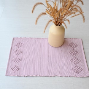 Pode incluir: Um tapete tecido rosa claro com um padrão de diamante nas bordas. O tapete está sobre um piso de madeira branca e um vaso com grama seca está no fundo.