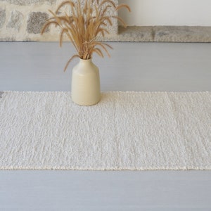 Puede incluir: Una alfombra blanca y esponjosa con flecos en los bordes. La alfombra est&aacute; colocada sobre un suelo gris claro con un jarr&oacute;n de hierba seca en el fondo.