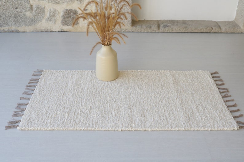 Puede incluir: Una alfombra blanca y esponjosa con flecos en los bordes. La alfombra est&aacute; colocada sobre un suelo gris claro con un jarr&oacute;n de hierba seca en el fondo.