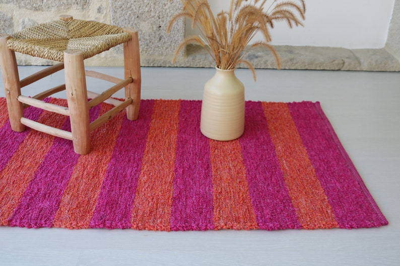 May include: A pink and orange striped rug with a wooden stool and a vase with dried flowers.